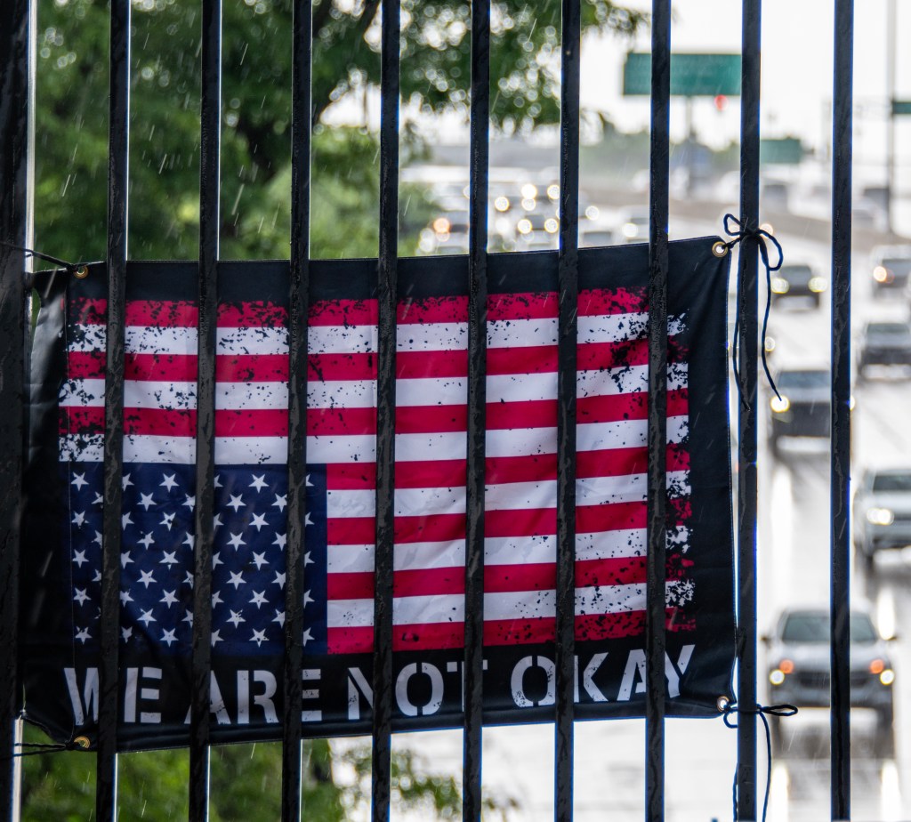 A distressed American flag with the phrase 'WE ARE NOT OKAY' displayed on a fence, with a busy road and traffic visible in the background.