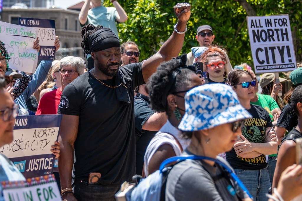 A diverse group of protesters gathering in a public space, holding signs advocating for social justice and community issues, with a focus on one person raising a fist in solidarity.