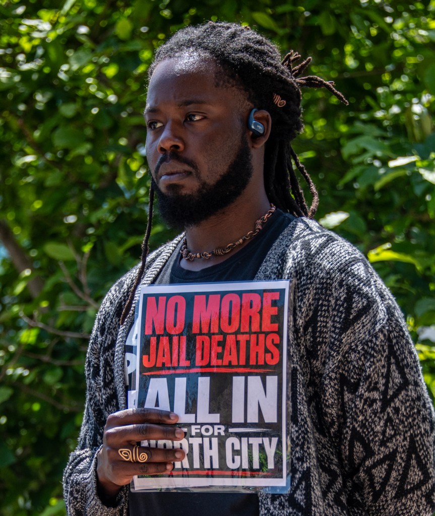 A person holding a sign that reads 'NO MORE JAIL DEATHS ALL IN FOR EARTH CITY' during a protest, surrounded by greenery.