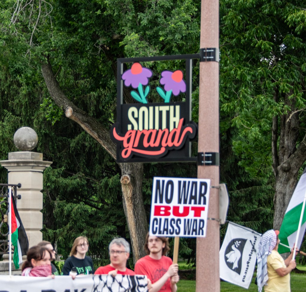 A diverse group of people gathered at a protest, holding banners and flags. A sign displaying 'South grand' with two flowers is visible in the background. One prominent sign reads 'NO WAR BUT CLASS WAR'.