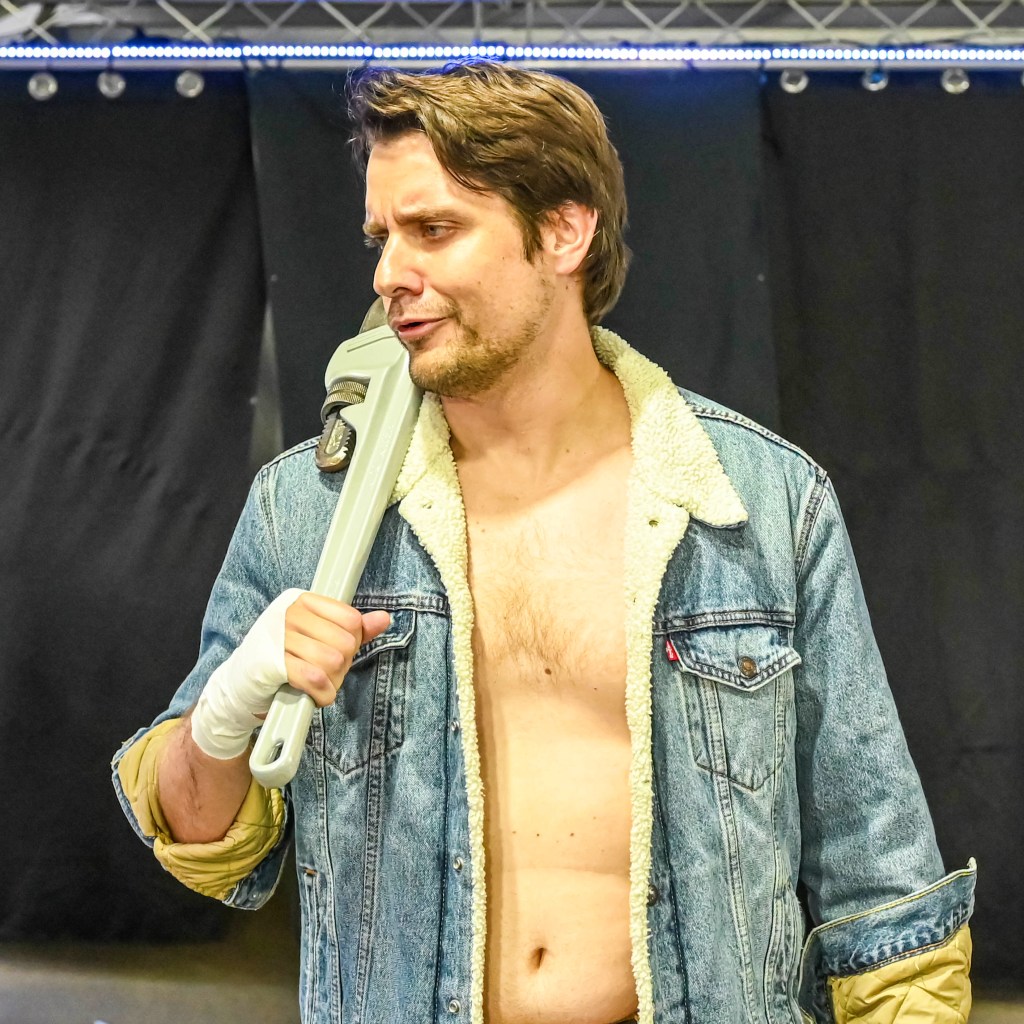 A man wearing a denim jacket with a shearling collar holds a large adjustable wrench on his shoulder, looking contemplative. He has a partially exposed chest and a bandaged hand, set against a dark background.