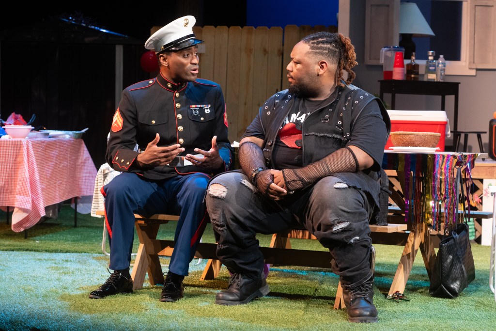 Two characters seated on a stage, one dressed in a military uniform and the other in casual punk attire, engaged in conversation. A picnic setting is visible in the background.