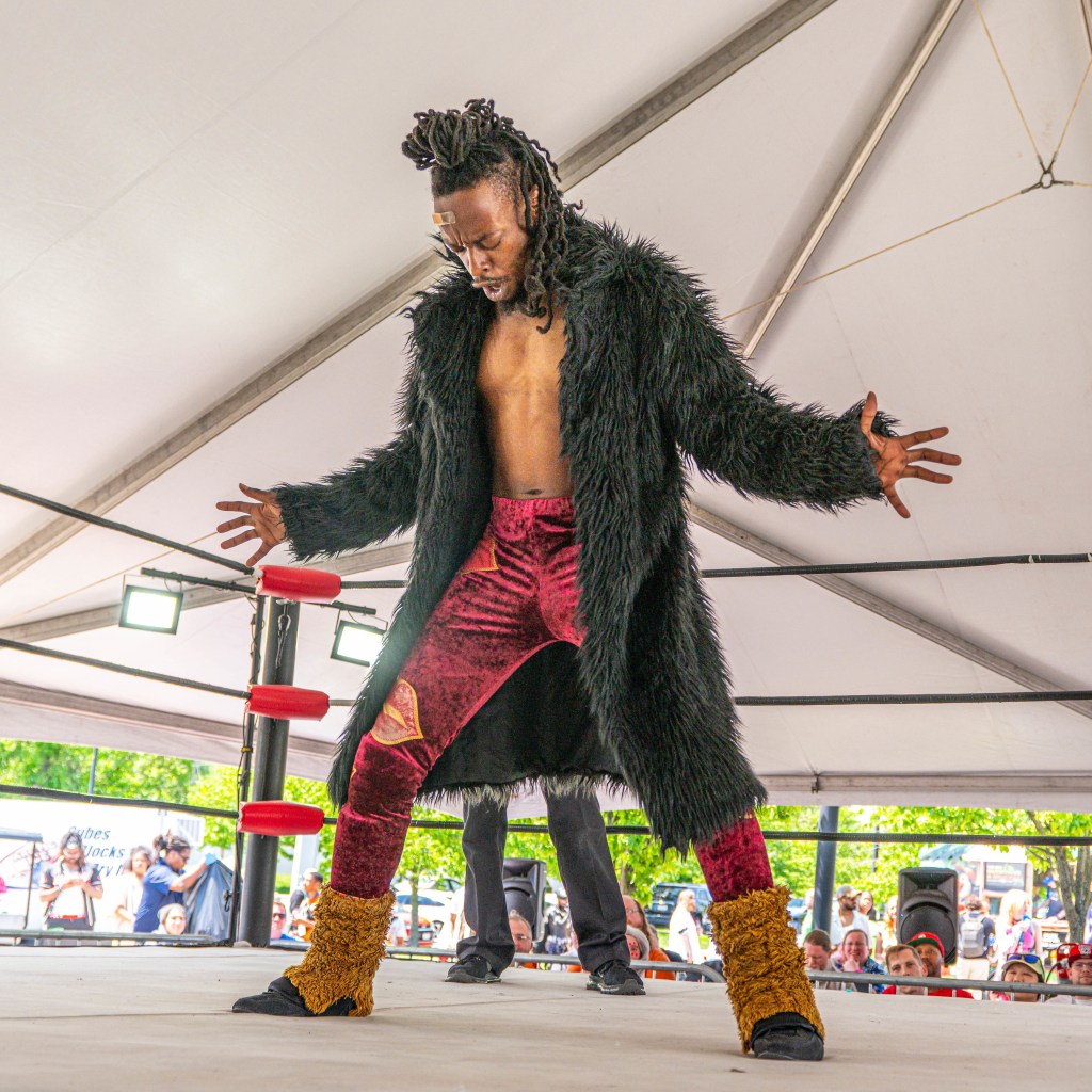 A professional wrestler dressed in a dramatic black shaggy coat and red pants poses on a wrestling stage, displaying a confident stance with outstretched arms.