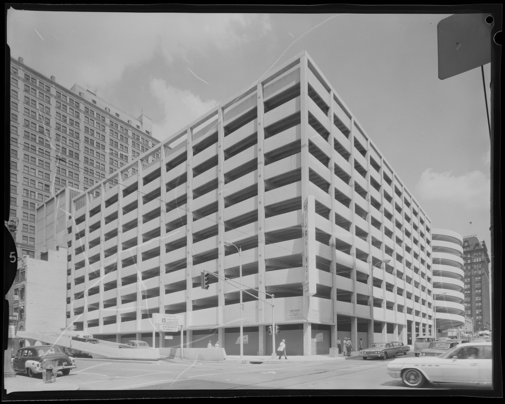 Black and white photograph of a multi-story parking garage with a modern concrete design, situated at an intersection with visible traffic and pedestrians nearby.