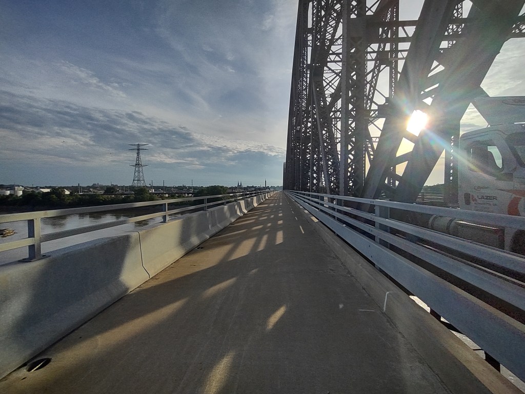 A view of a bridge walkway with metal beams and structures, sunlight shining through, and a river below.