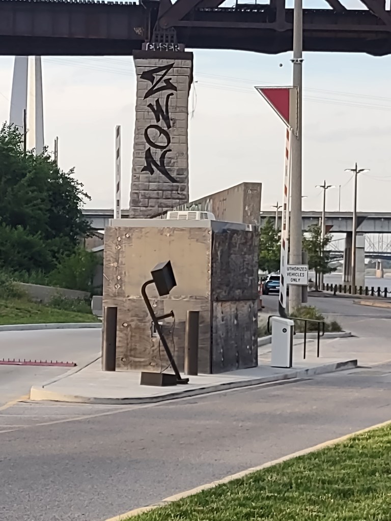 A weathered concrete structure with graffiti and a sleek, modern signpost nearby, surrounded by greenery and paved roadways.