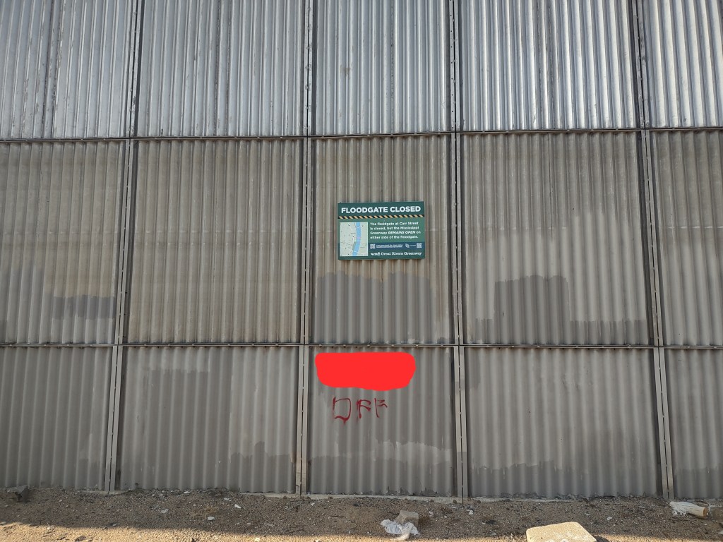 A corrugated metal wall with a sign reading 'FLOODGATE CLOSED' mounted at the top. Below the sign, there is a red smudge or graffiti covering part of the wall.
