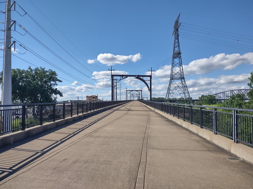 A wide, empty pathway on a bridge with metal railings, surrounded by greenery and power lines under a clear blue sky with fluffy clouds.