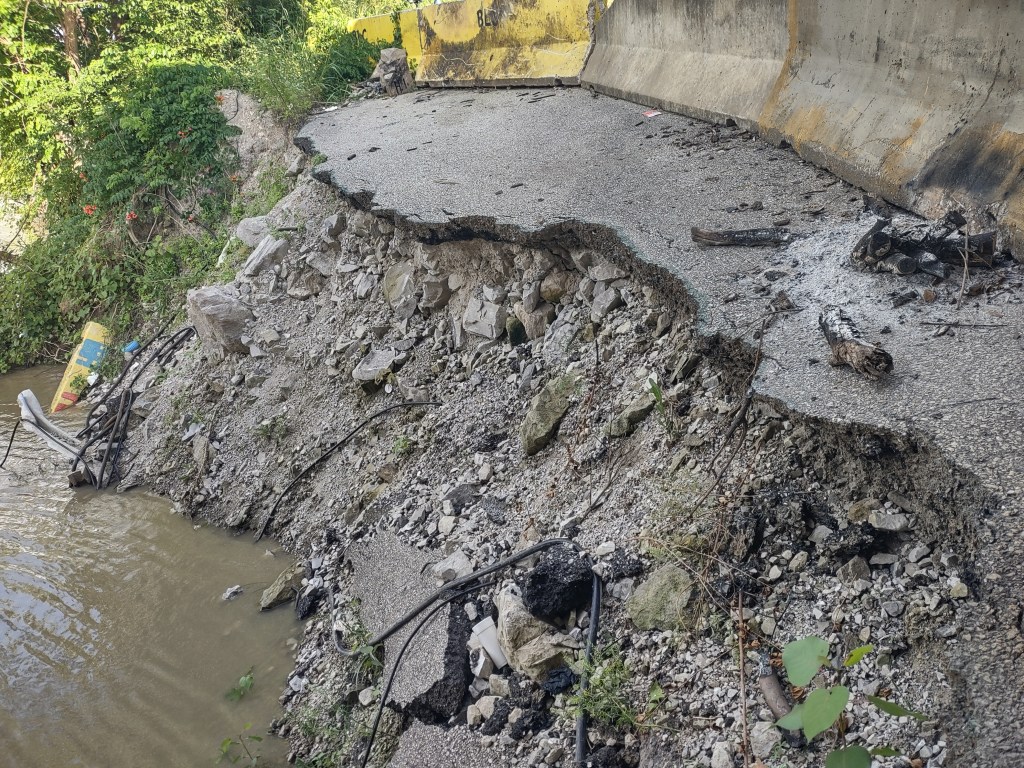 Eroded roadway edge with exposed gravel and debris near a water body, showcasing signs of damage and disrepair.