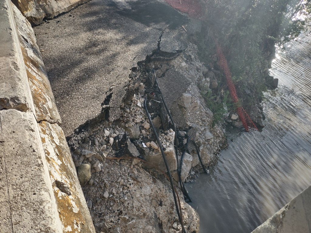 A section of cracked and damaged pavement next to a body of water, with exposed rocks and a black utility line visible in the debris.