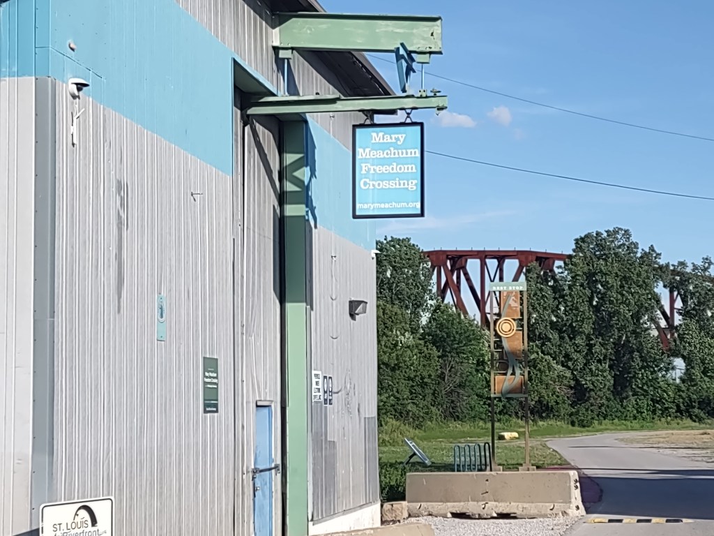 Exterior view of the Mary Meachum Freedom Crossing sign mounted on a building, with a bridge and vegetation in the background.
