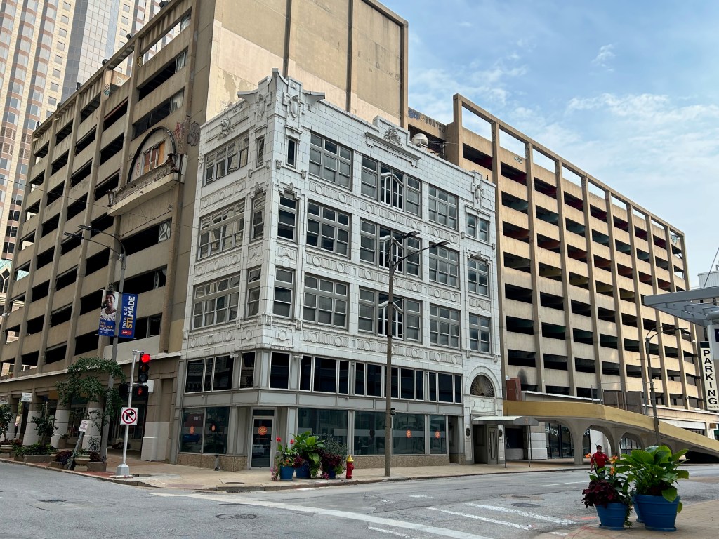 A historic white building with ornate architectural details stands next to a modern parking garage in an urban setting. The street is lined with potted plants, and a traffic light is visible.