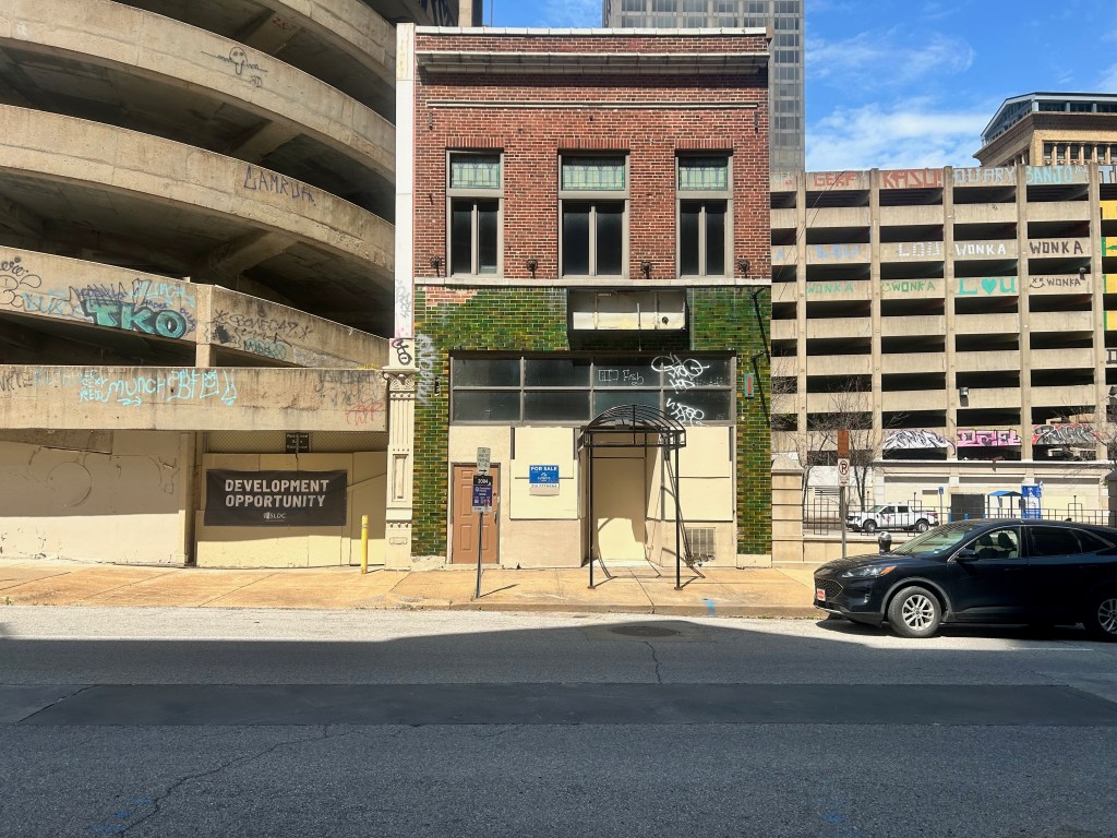 Abandoned building with green tiles and a sign reading 'Development Opportunity' next to a multi-story parking garage, under a bright blue sky.