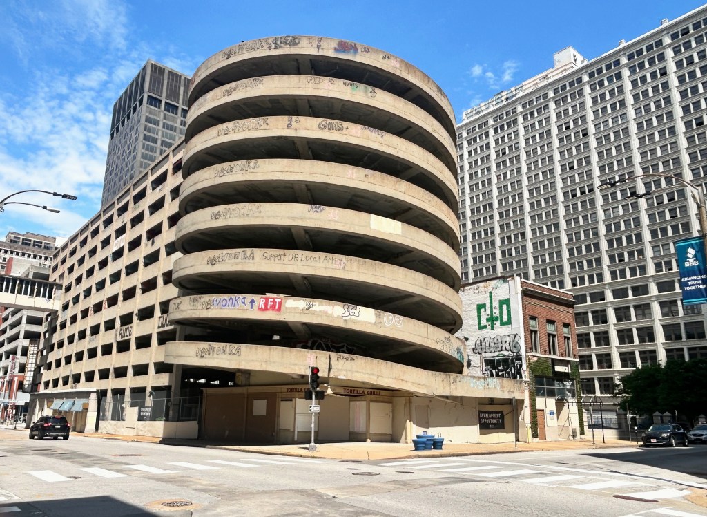 An abandoned circular parking garage with graffiti on its walls, located in an urban area, surrounded by taller buildings and a clear blue sky.