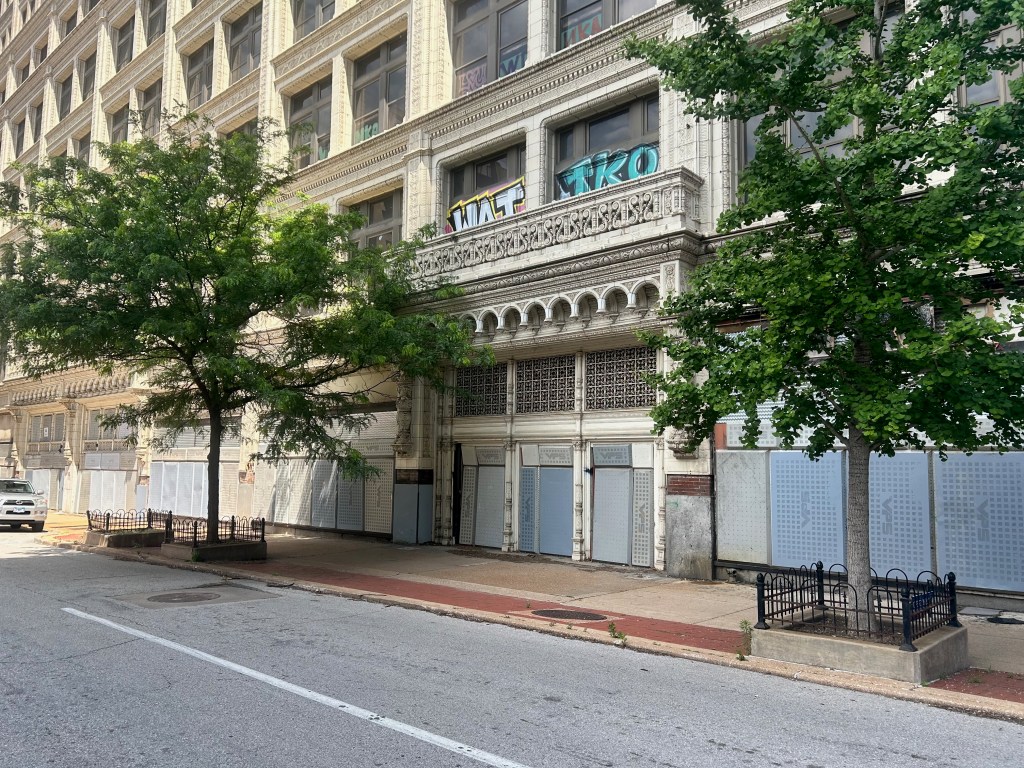 Abandoned building facade with boarded windows and trees lining the street.