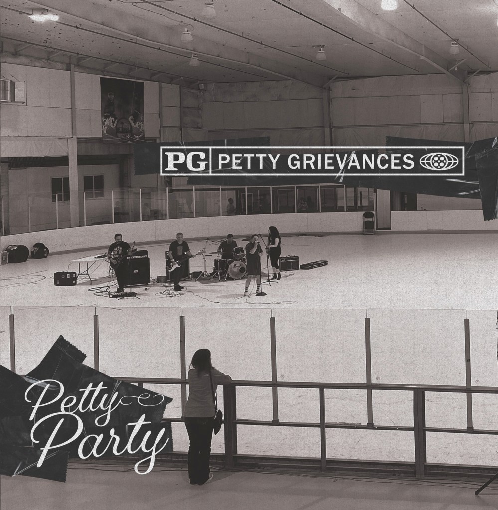 A black and white image of a band performing on an ice rink, with a vocal artist and musicians on stage. A person stands in the foreground watching the performance, and a large banner displaying 'Petty Grievances' is visible above the stage.