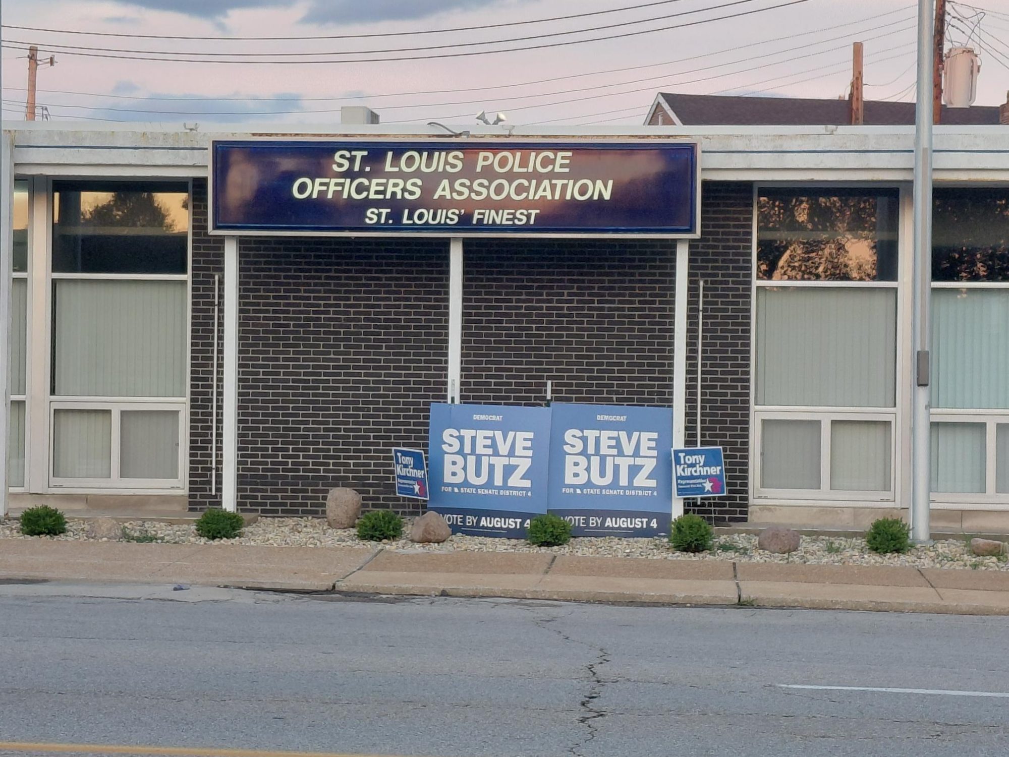 Exterior view of the St. Louis Police Officers Association building with a sign above that reads 'ST. LOUIS POLICE OFFICERS ASSOCIATION ST. LOUIS' FINEST'. In front, there are campaign signs for Steve Butz and Tony Kirchner.