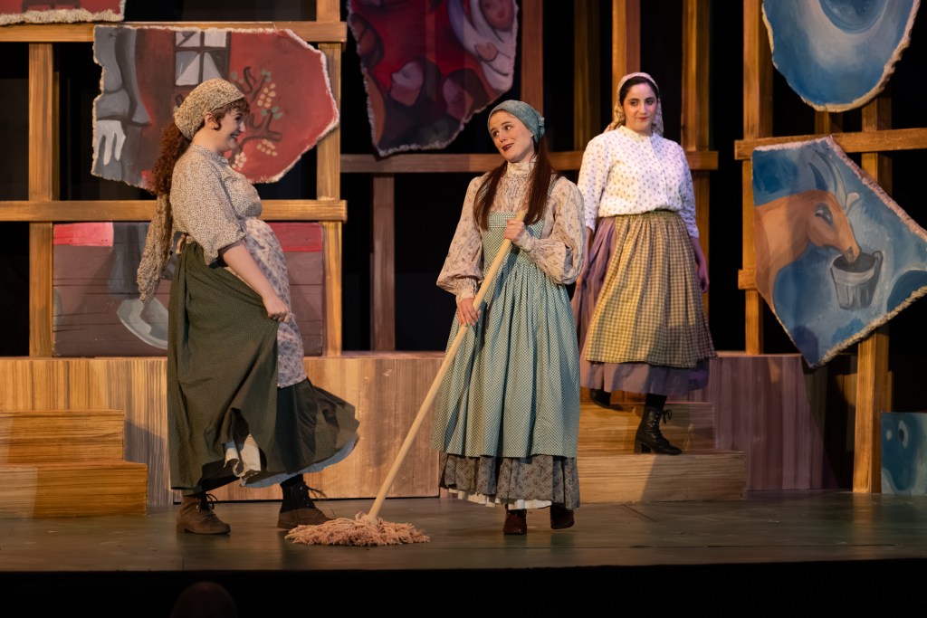 Three women performing on stage in period costumes, with the central character holding a mop and engaging with the others.