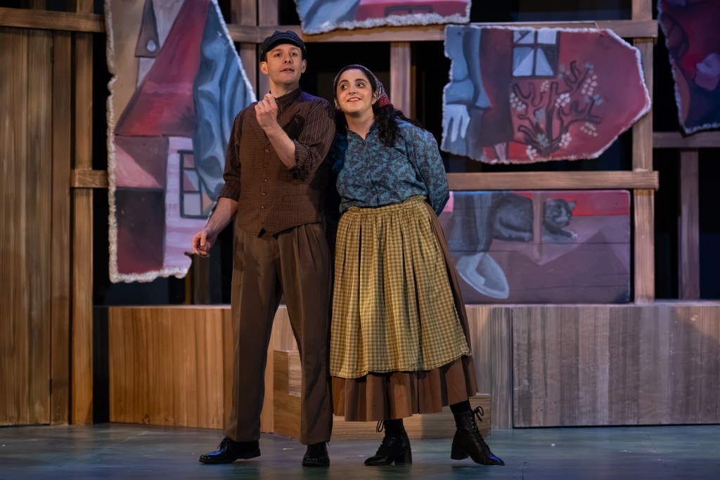 Two actors performing in a theatrical scene, standing together in front of painted stage backdrops. One actor is wearing a hat and a brown vest, while the other is in a blue blouse and a long plaid skirt.