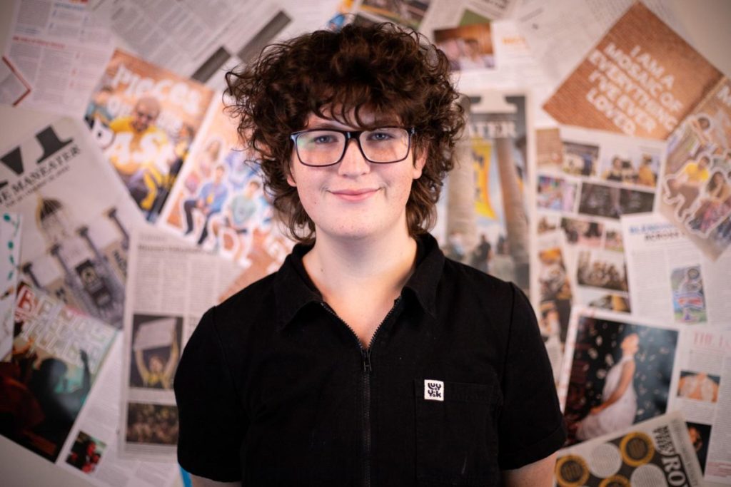 A person with curly hair and glasses stands in front of a collage of newspaper clippings and photos.