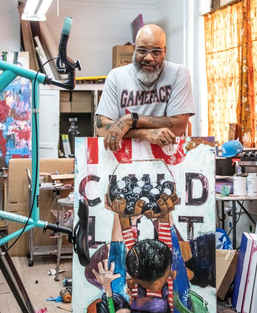 A bearded man wearing glasses and a 'GAMEFACE' t-shirt stands in an art studio, resting his arms on a colorful artwork featuring raised hands holding black balls with numbers. The studio has various art supplies and colorful decorations in the background.