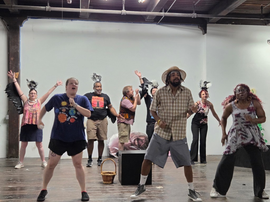 A diverse group of people performing in an art space, wearing casual attire and showing lively expressions. Some participants have costumes with wings, while others are engaged in energetic movements.