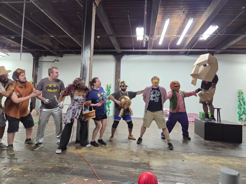 A group of eight actors in costumes performing a scene in an indoor space, with various props and a playful atmosphere. Some actors are wearing masks or themed outfits, while others hold props like a basket and a shield.