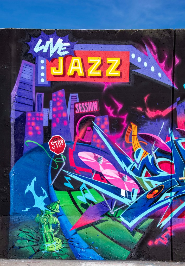 Colorful graffiti mural featuring the words 'Live Jazz' and 'Session', with vibrant shapes and musical elements including turntables and city skyline in the background.