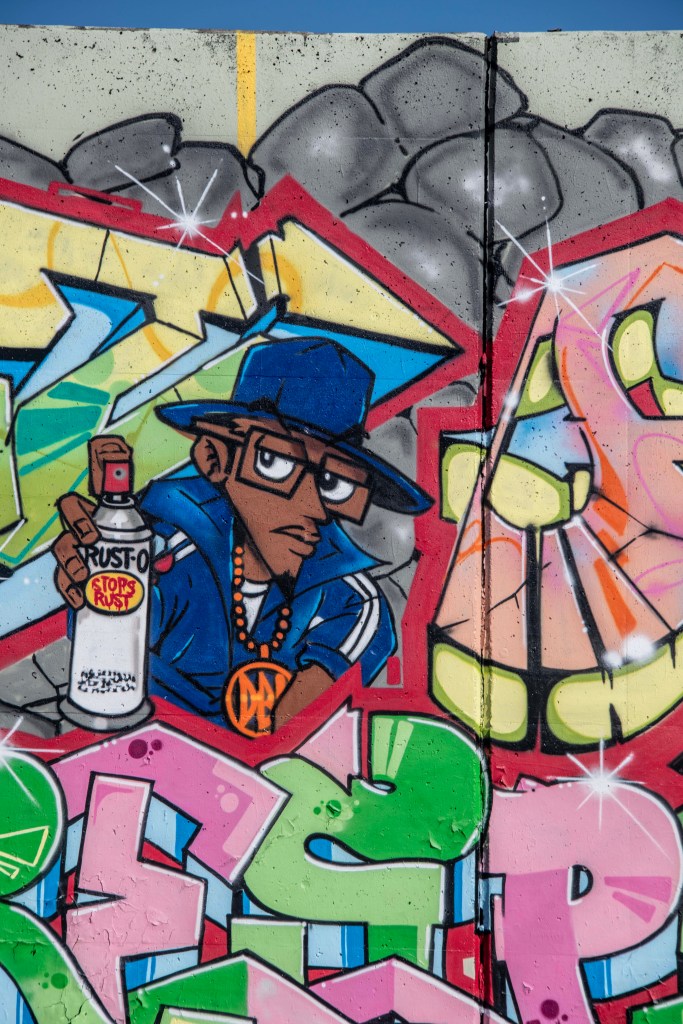 A colorful mural featuring a cartoon character wearing a blue hat and glasses, holding a can of Rust-Oleum spray paint. The background showcases vibrant graffiti art with various shapes and colors.
