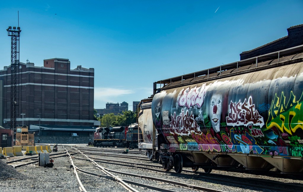 A train yard scene featuring graffiti-covered train cars, with a brick building in the background and clear blue skies.