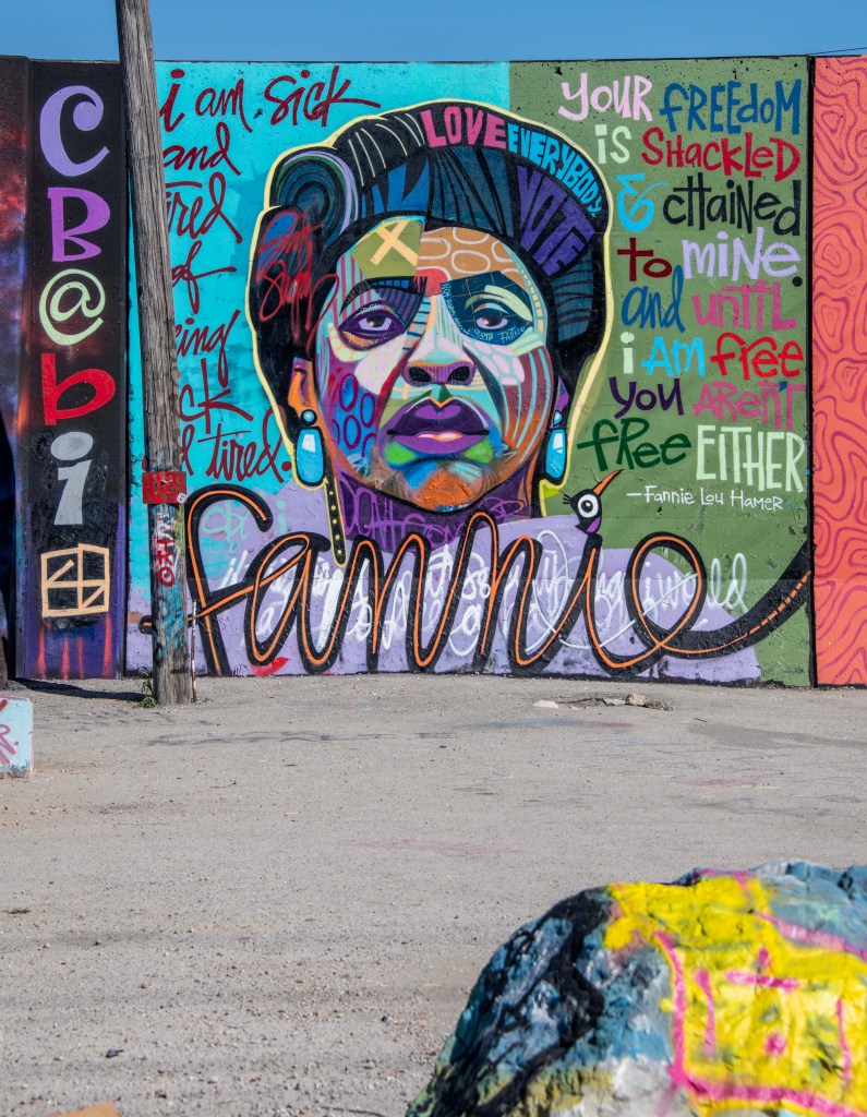 A vibrant mural depicting a woman with dark hair and bold features, surrounded by colorful graffiti phrases emphasizing freedom and equality. The word 'Fannie' is prominently featured, referencing Fannie Lou Hamer, along with various artistic symbols and text elements.