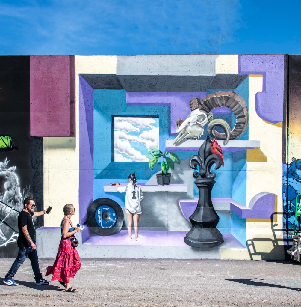 A vibrant mural featuring a woman reaching out from a stylized interior scene, surrounded by colorful geometric shapes, a plant, and a ram skull, with pedestrians walking in front.