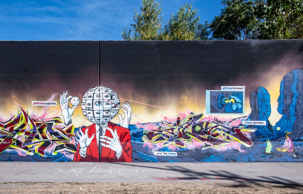 A colorful mural depicting a figure with a globe-like head covered in eyes, dressed in a red suit, surrounded by graffiti art. The mural includes speech bubbles referencing topics such as 'Material Refinement' and 'Hide the Source'.