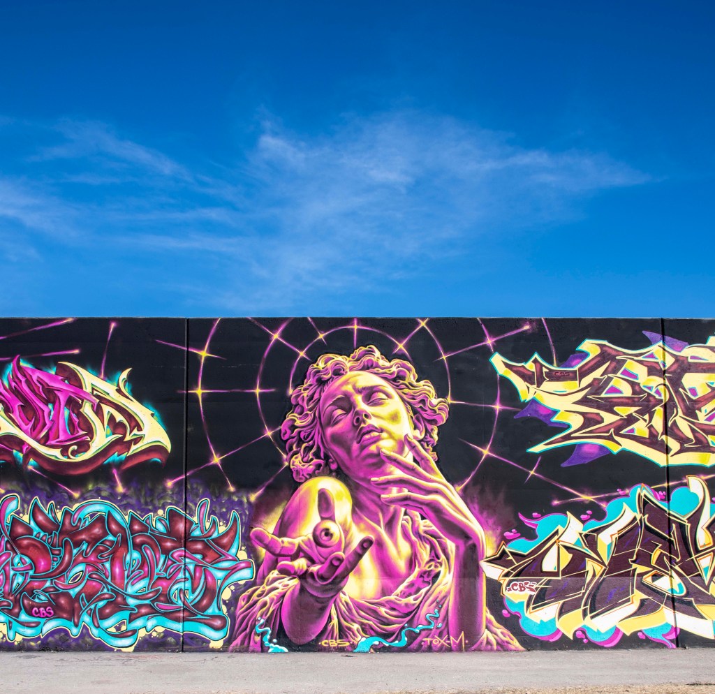 A vibrant mural featuring a stylized figure with flowing hair, reaching out with one hand surrounded by colorful graffiti elements and a blue sky background.