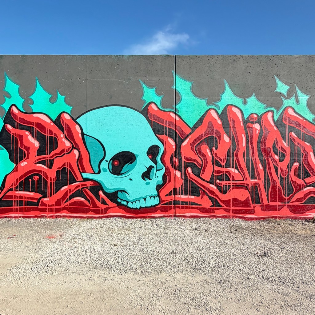 Colorful graffiti featuring a large turquoise skull with red details and bold lettering, set against a grey wall with a blue sky background.