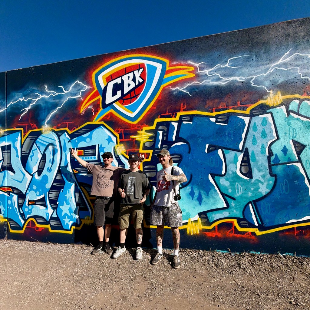 Three individuals posing in front of a colorful graffiti wall featuring the letters 'CBK' and vibrant designs.