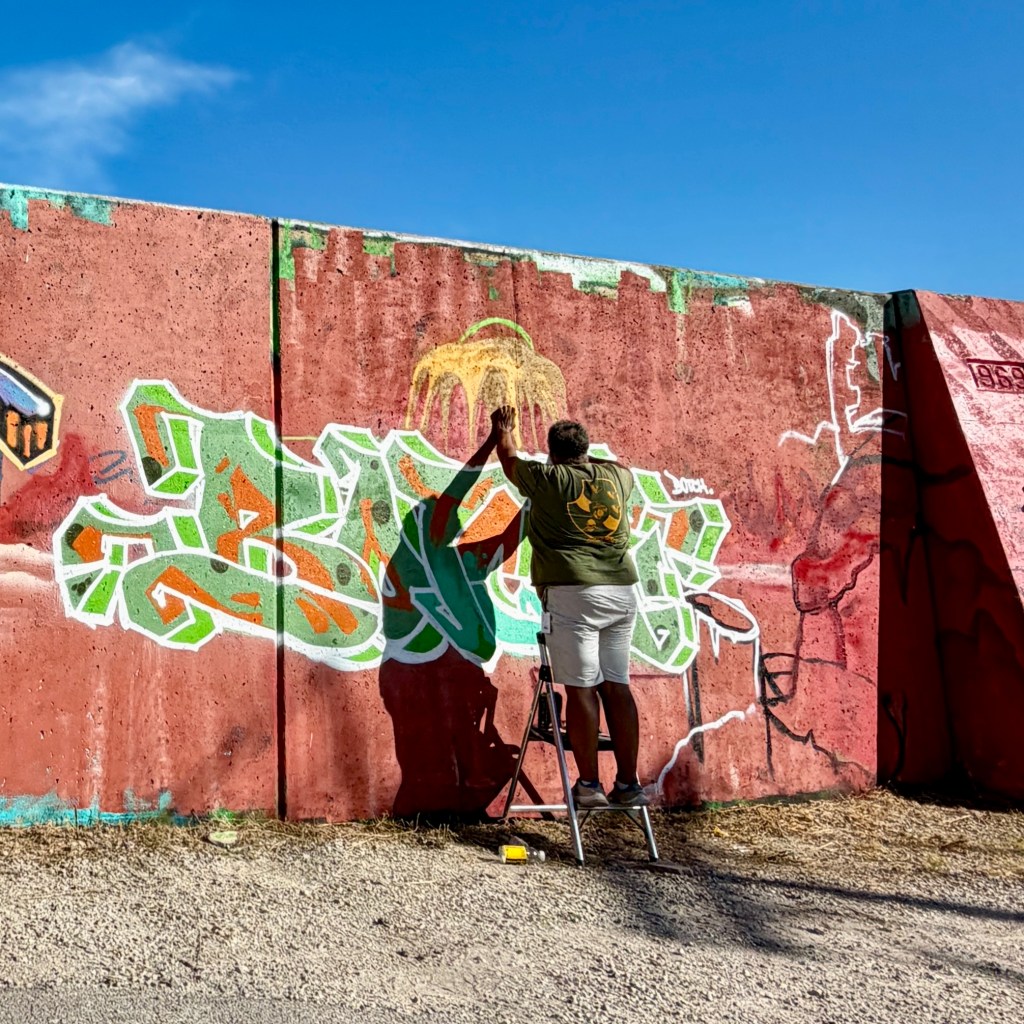 A person painting graffiti on a pink concrete wall while standing on a ladder, with blue sky in the background.