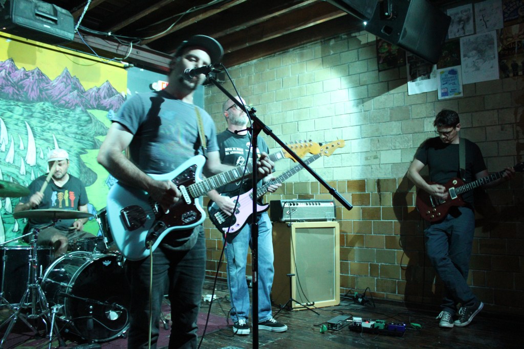 A band performing live on stage, featuring four musicians. One is playing drums, while the others are playing electric guitars. The backdrop is decorated with a colorful mural of mountains and abstract shapes. The venue has a casual atmosphere with dim lighting.
