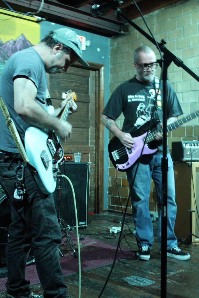 Two musicians performing on stage with electric guitars. One man is playing a white guitar while the other is playing a purple bass guitar.