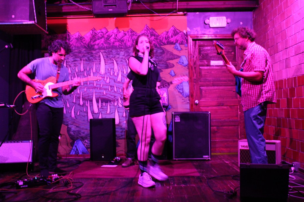 A live band performing on stage in a colorful venue with a mural in the background. The vocalist is singing while two guitarists play instruments, and a drummer is partially visible.