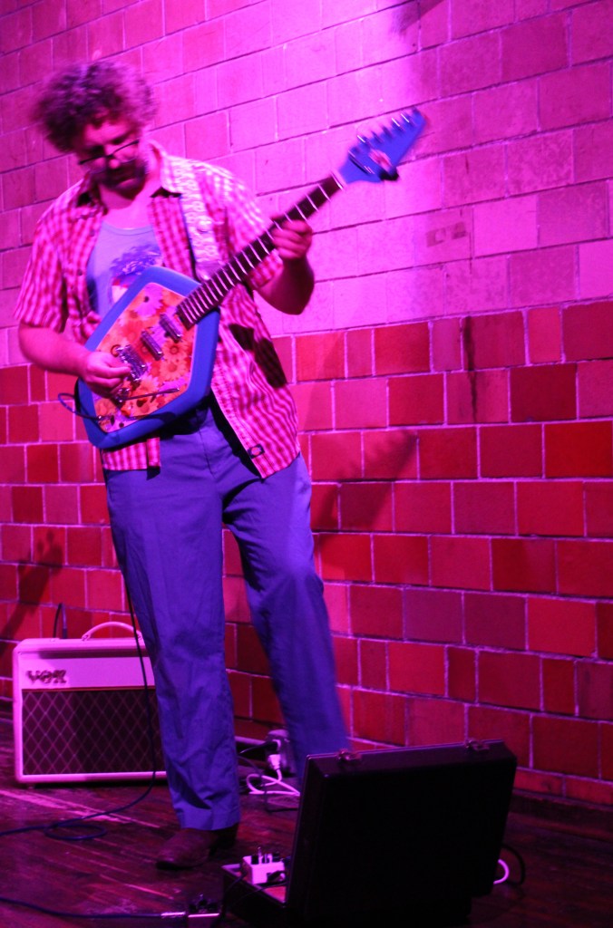 A musician playing a unique blue guitar on stage, wearing a plaid shirt and blue pants, with a brick wall in the background illuminated by purple lighting.