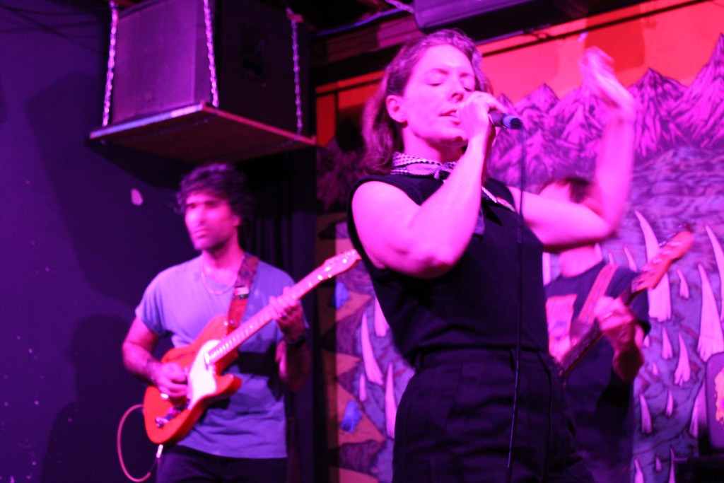 A female singer passionately performing on stage with a microphone, while a male guitarist plays in the background. The setting includes colorful artwork on the wall and dramatic lighting effects.