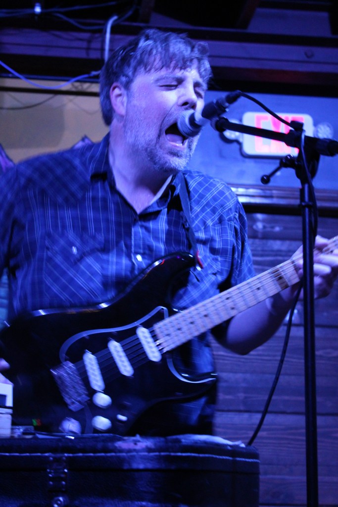 A man passionately singing into a microphone while playing an electric guitar on stage, with a blurry background and dim lighting.