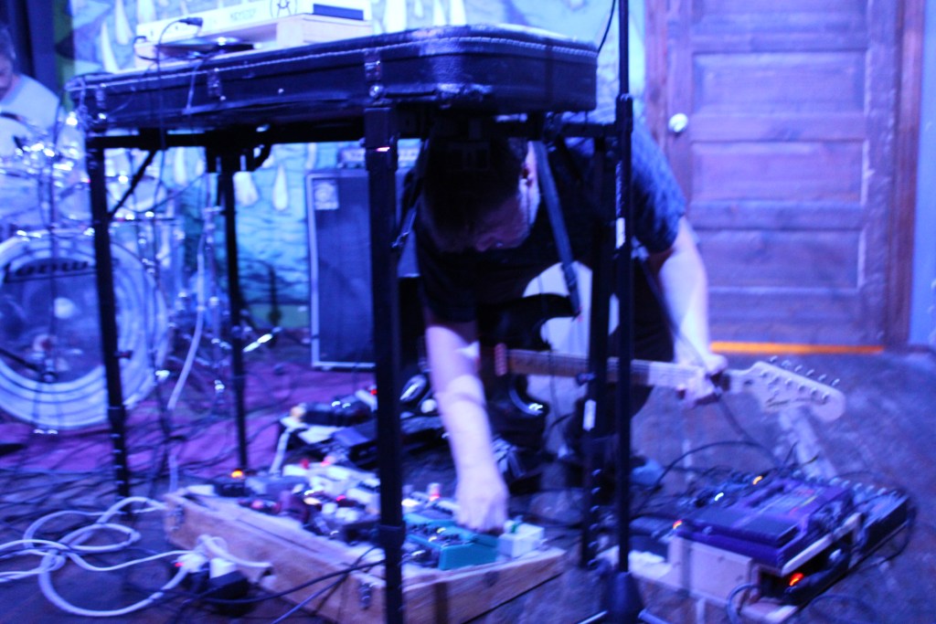 Musician adjusting effects pedals while playing an electric guitar on stage, with a drum set visible in the background.
