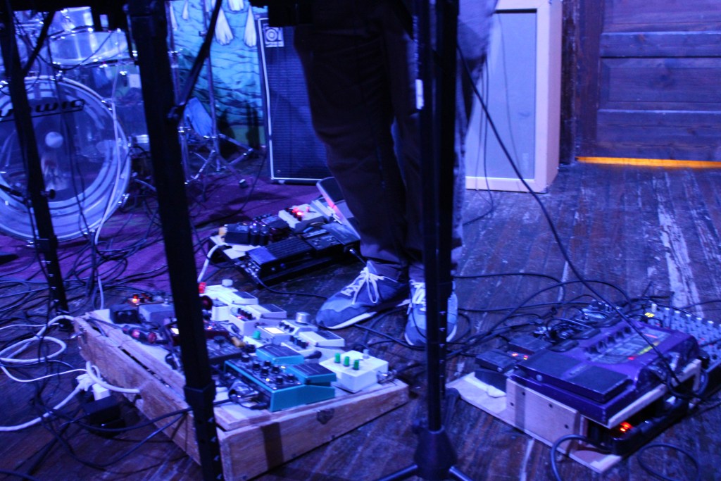 A musician's feet standing on various guitar pedals on a stage, with a drumset and amplifiers in the background.