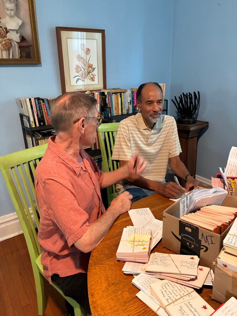 Two men engaged in conversation and writing at a wooden table surrounded by bookshelves and artwork, with stacks of notes and postcards visible.