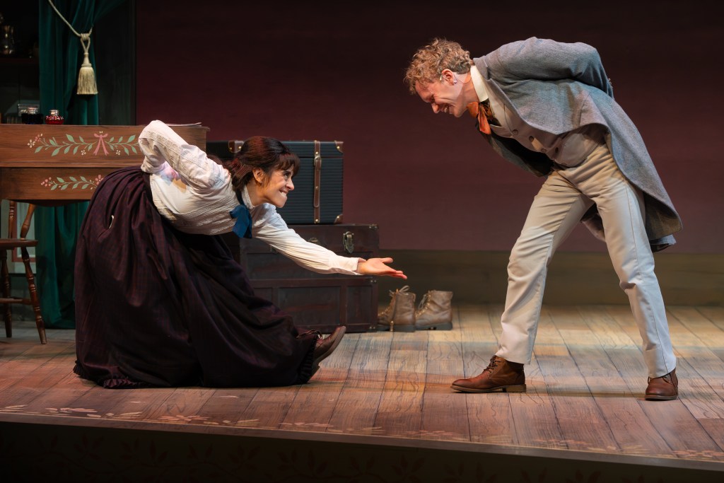 A man and a woman perform on stage, with the woman bending down and extending her hand while the man leans forward, creating a playful interaction. They are dressed in period costumes, and the stage has a rustic wooden floor and background props.
