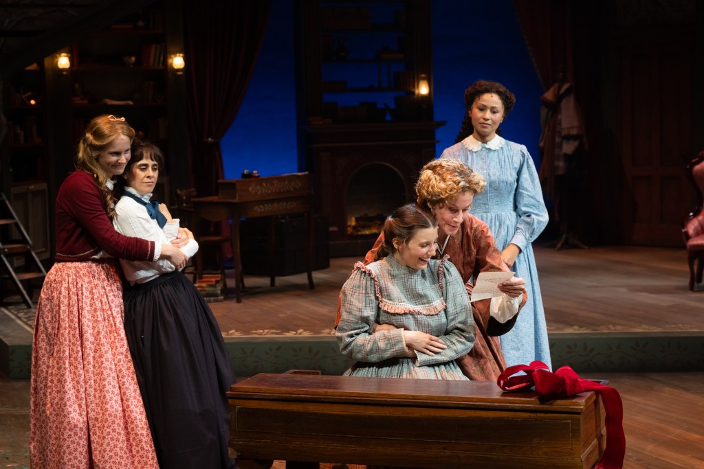 A scene from a theatrical performance featuring four women in historical costumes, gathered around a table. One woman sits reading a letter, while another leans affectionately on a friend. The background features a cozy, warmly lit room with period-appropriate decor.