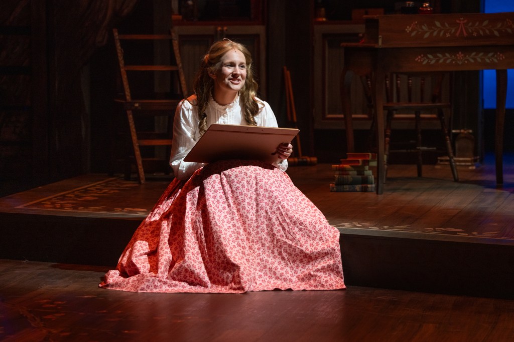A young woman in a white blouse and a red-patterned skirt sits on a stage, holding a sketchbook and smiling. The background features wooden furniture and shelves, creating a theatrical setting.