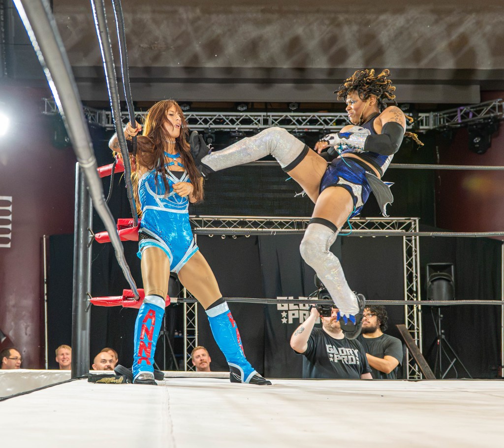 A female wrestler performs a kick on her opponent during a wrestling match in a ring, with spectators in the background.